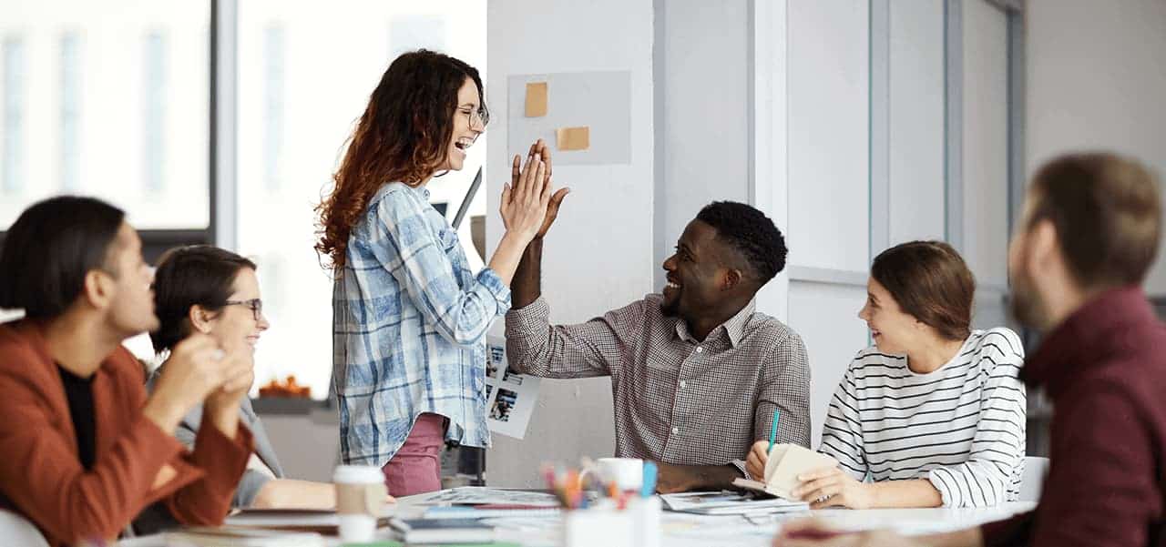 Eine Frau steht an einem Tisch, an dem eine Gruppe Menschen sitzen und gibt einem Mann ein High Five